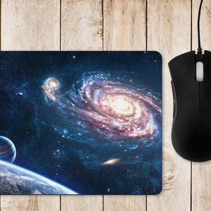 May include: A mouse mat with a space-themed design featuring a swirling galaxy with pink, orange, and yellow hues against a dark blue background with scattered stars. A small planet is visible in the bottom left corner.