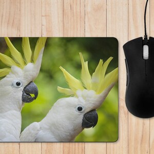 May include: Mouse mat with a colourful image of two white cockatoos with yellow crests and black beaks. The background is a blurred green. A black computer mouse is positioned to the right of the mouse mat.