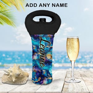 May include: A black wine bottle carrier with a handle, displaying a vibrant blue, purple, and gold abstract design. The name "Leanne" is written in gold script. A glass of champagne and a seashell are also present.