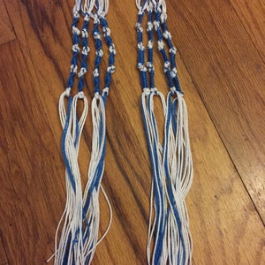 Peut inclure: Deux longues mèches de ficelle bleue et blanche sont nouées ensemble dans un motif de nœuds. Les ficelles pendent et ont une frange au bas.