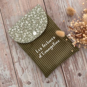 Peut inclure: Une pochette à livre en velours côtelé vert olive avec un rabat à motif floral blanc et un bouton-pression blanc. L'avant présente le texte cursif blanc Les lectures d'Evangeline. Une petite étiquette blanche est visible sur le côté.