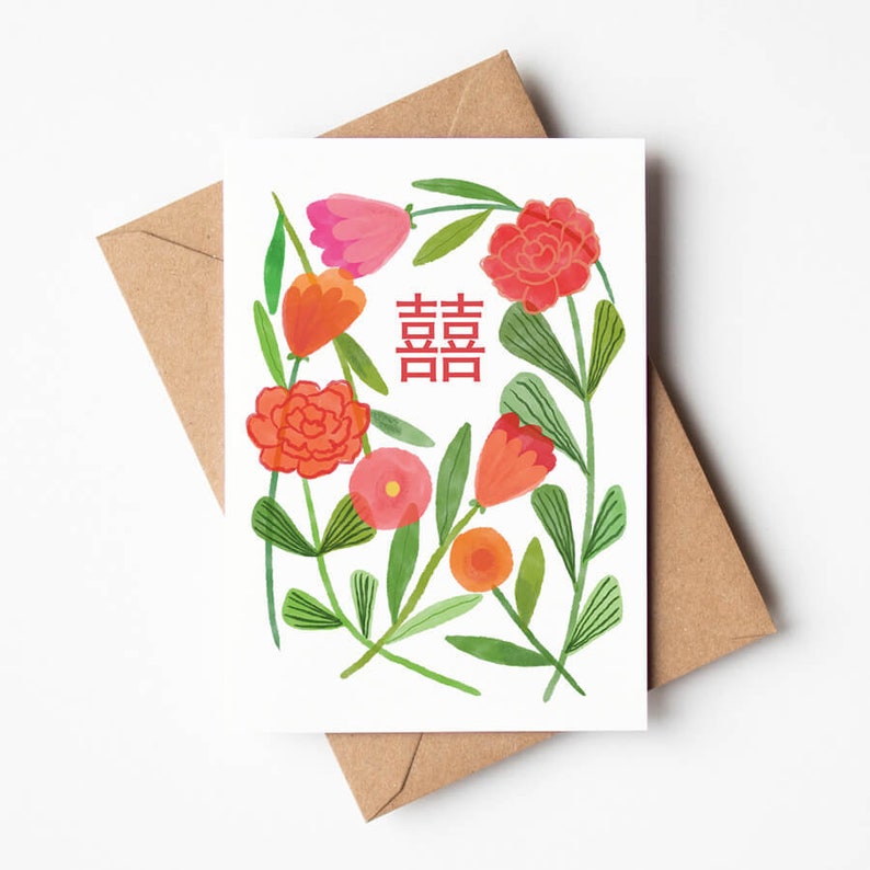 Puede incluir: Una tarjeta de felicitaci&oacute;n con un dise&ntilde;o floral con flores rojas, rosas y naranjas, hojas verdes y el car&aacute;cter chino para la doble felicidad. La tarjeta est&aacute; sobre un fondo blanco, con un sobre marr&oacute;n detr&aacute;s.