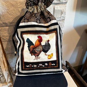 May include: A drawstring bag featuring a rooster and hen illustration with two chicks. The bag has a black and white striped border and a floral patterned top. The bag is sitting on a dark blue wooden stool.