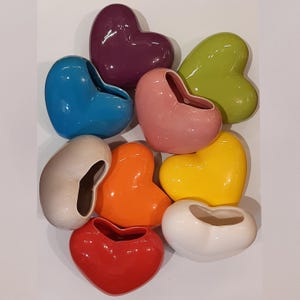 Può includere: Un set di nove vasi in ceramica a forma di cuore in vari colori, tra cui viola, blu, rosa, beige, arancione, giallo, rosso e bianco.