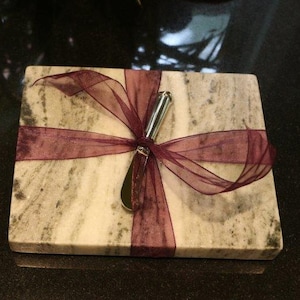 May include: A white marble cheese board with a silver cheese knife tied with a burgundy ribbon.