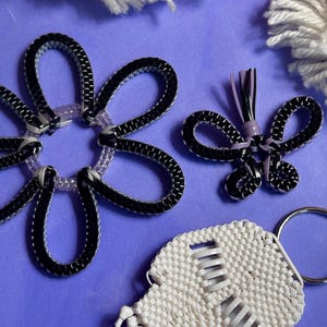May include: A collection of handmade accessories, including a black and white flower, a black and purple bow, and a white skull-shaped keychain. The items are set against a purple background, with white yarn accents.