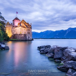 May include: A medieval castle with a flag on top, illuminated at dusk, sits on a rocky shore overlooking a calm lake with mountains in the background.