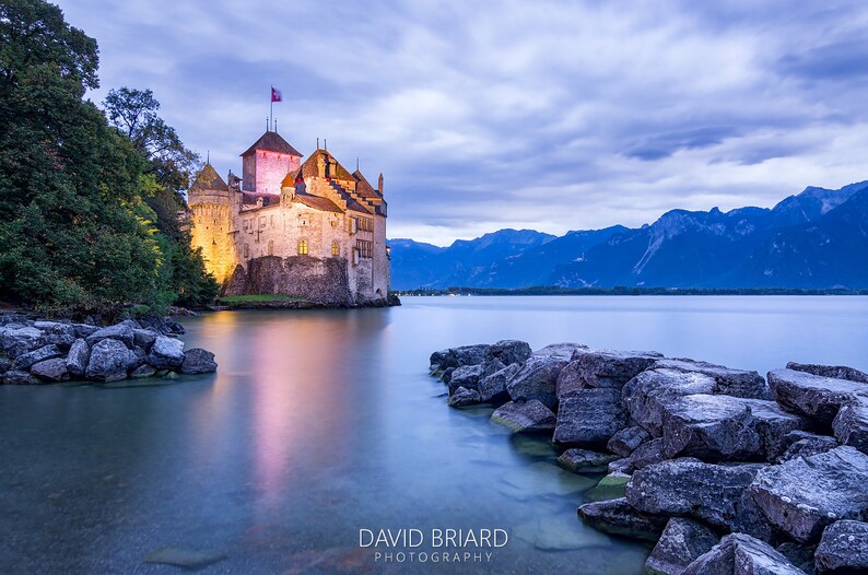 May include: A medieval castle with a flag on top, illuminated at dusk, sits on a rocky shore overlooking a calm lake with mountains in the background.