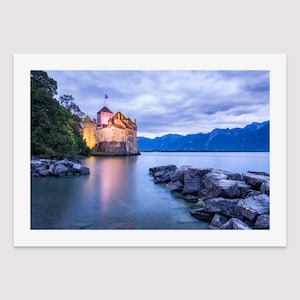 May include: A stone castle with a flag on top sits on the edge of a lake with mountains in the background. The water is calm and reflects the castle and the sky. The sky is a light blue with some clouds.