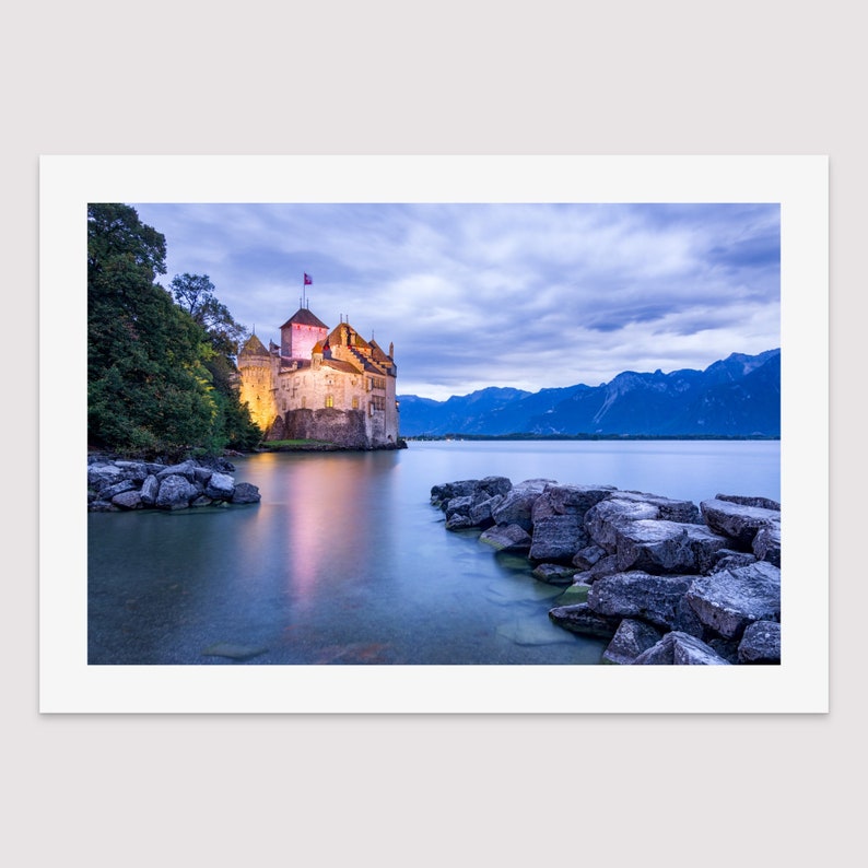 May include: A stone castle with a flag on top sits on the edge of a lake with mountains in the background. The water is calm and reflects the castle and the sky. The sky is a light blue with some clouds.