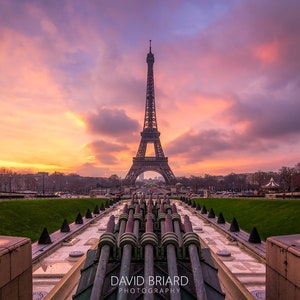 Puede incluir: La Torre Eiffel en Par&iacute;s, Francia, se alza contra un cielo de atardecer vibrante con tonos naranja, rosa y p&uacute;rpura. En primer plano, una fuente con tuber&iacute;as met&aacute;licas. La imagen incluye el texto "DAVID BRIARD PHOTOGRAPHY".