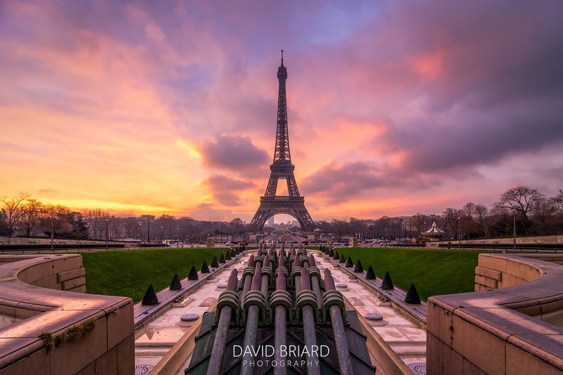 Puede incluir: La Torre Eiffel en Par&iacute;s, Francia, se alza contra un cielo de atardecer vibrante con tonos naranja, rosa y p&uacute;rpura. En primer plano, una fuente con tuber&iacute;as met&aacute;licas. La imagen incluye el texto "DAVID BRIARD PHOTOGRAPHY".