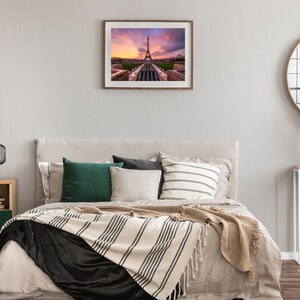 Puede incluir: Una impresi&oacute;n enmarcada de la Torre Eiffel al atardecer cuelga sobre una cama con almohadas y una manta a rayas. Una mesita de noche con una l&aacute;mpara y un espejo redondo completan la decoraci&oacute;n del dormitorio.