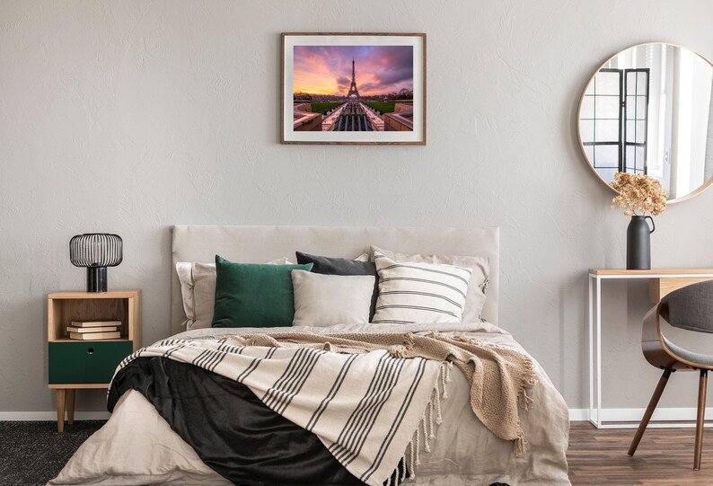 Puede incluir: Una impresi&oacute;n enmarcada de la Torre Eiffel al atardecer cuelga sobre una cama con almohadas y una manta a rayas. Una mesita de noche con una l&aacute;mpara y un espejo redondo completan la decoraci&oacute;n del dormitorio.