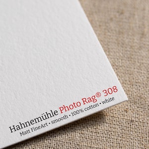 May include: Close-up of a white Hahnem&uuml;hle Photo Rag 308 paper sample. The text reads "Matt FineArt - smooth - 100% cotton - white".