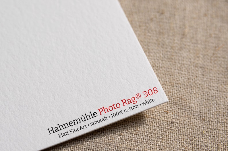 Puede incluir: Primer plano de una hoja blanca de papel Hahnem&uuml;hle Photo Rag 308. El papel es liso y est&aacute; hecho de 100% algod&oacute;n. El texto "Hahnem&uuml;hle Photo Rag 308" est&aacute; impreso en rojo, con texto adicional en negro.