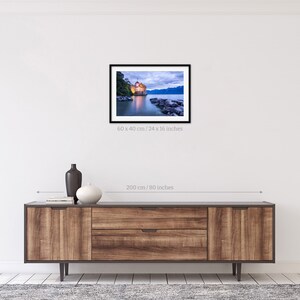 May include: A photograph of a castle on a lake at dusk. The castle is lit up and the water is reflecting the light. The sky is a soft blue and there are clouds in the sky. The image is 24 inches by 16 inches.