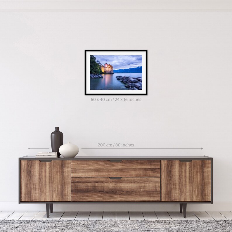 May include: A photograph of a castle on a lake at dusk. The castle is lit up and the water is reflecting the light. The sky is a soft blue and there are clouds in the sky. The image is 24 inches by 16 inches.