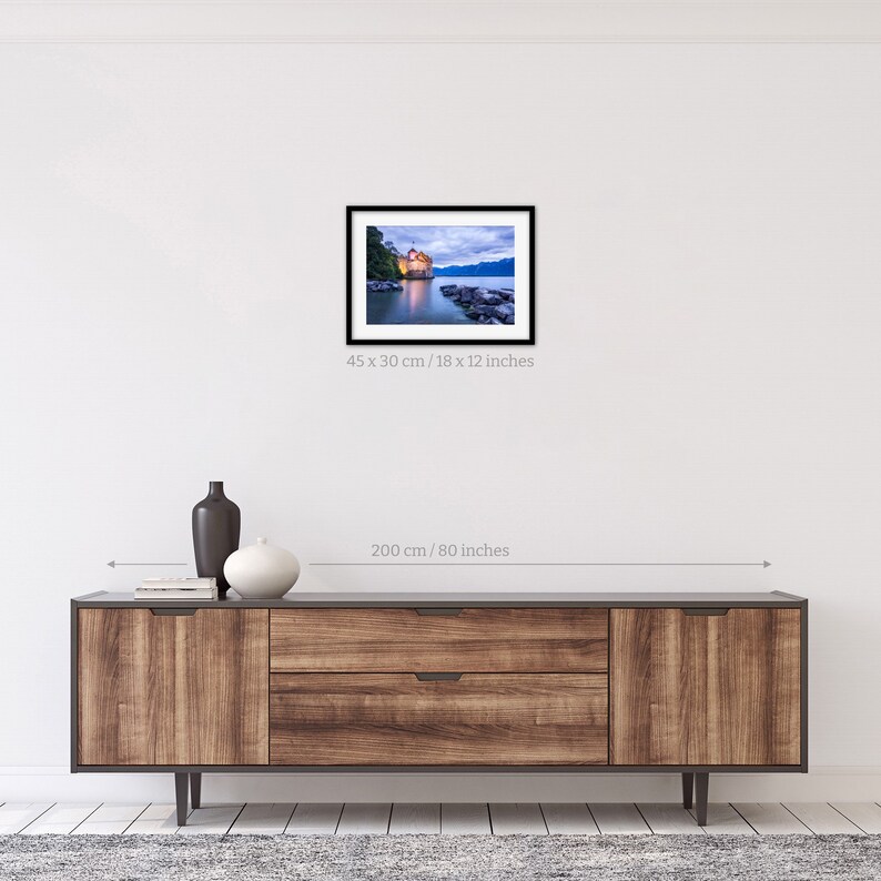 May include: A framed photograph of a castle on a lake at dusk. The castle is lit up and reflected in the water. The sky is a soft blue with clouds. The image is 18 inches by 12 inches.