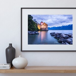 May include: A framed photograph of a castle on a lake at dusk. The castle is lit up and there are mountains in the background. The water is calm and reflects the light from the castle.