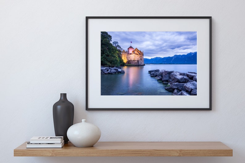 May include: A framed photograph of a castle on a lake at dusk. The castle is lit up and there are mountains in the background. The water is calm and reflects the light from the castle.