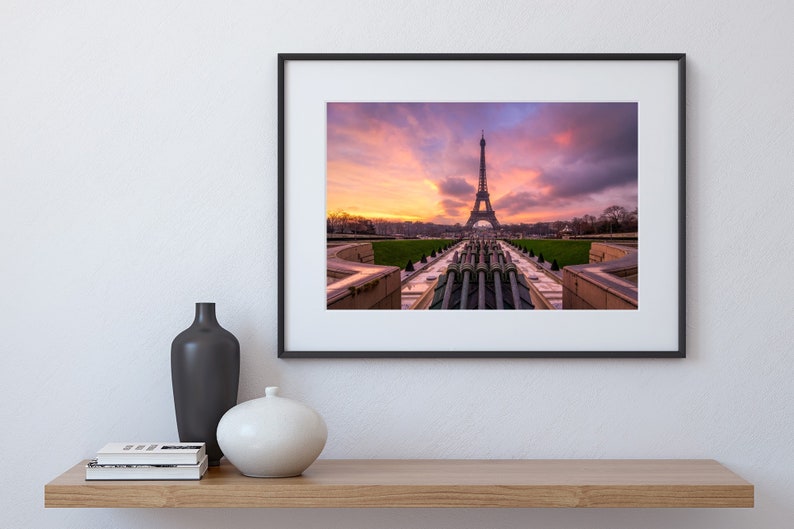 Puede incluir: Impresi&oacute;n enmarcada de la Torre Eiffel al atardecer. La ic&oacute;nica torre se alza sobre un cielo vibrante de tonos naranja, rosa y p&uacute;rpura. La imagen est&aacute; enmarcada en negro y se exhibe en una estanter&iacute;a de madera con jarrones.