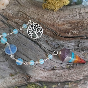 Puede incluir: Un p&eacute;ndulo de plata con un dije de &aacute;rbol de la vida y una punta de cristal multicolor. El p&eacute;ndulo est&aacute; encordado con cuentas de color azul claro y cuelga de una cadena de plata.