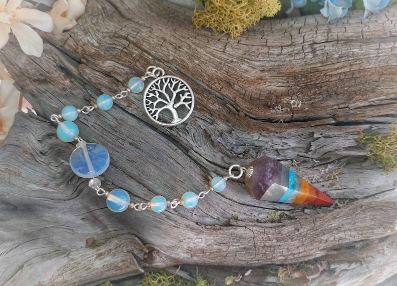 Puede incluir: Un p&eacute;ndulo de plata con un dije de &aacute;rbol de la vida y una punta de cristal multicolor. El p&eacute;ndulo est&aacute; encordado con cuentas de color azul claro y cuelga de una cadena de plata.