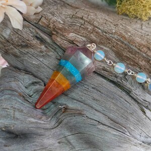 Puede incluir: Un p&eacute;ndulo de cristal arco&iacute;ris con una cadena de plata y cuentas de &oacute;palo blanco. El p&eacute;ndulo es un cristal puntiagudo con capas de rojo, naranja, amarillo, azul y morado.