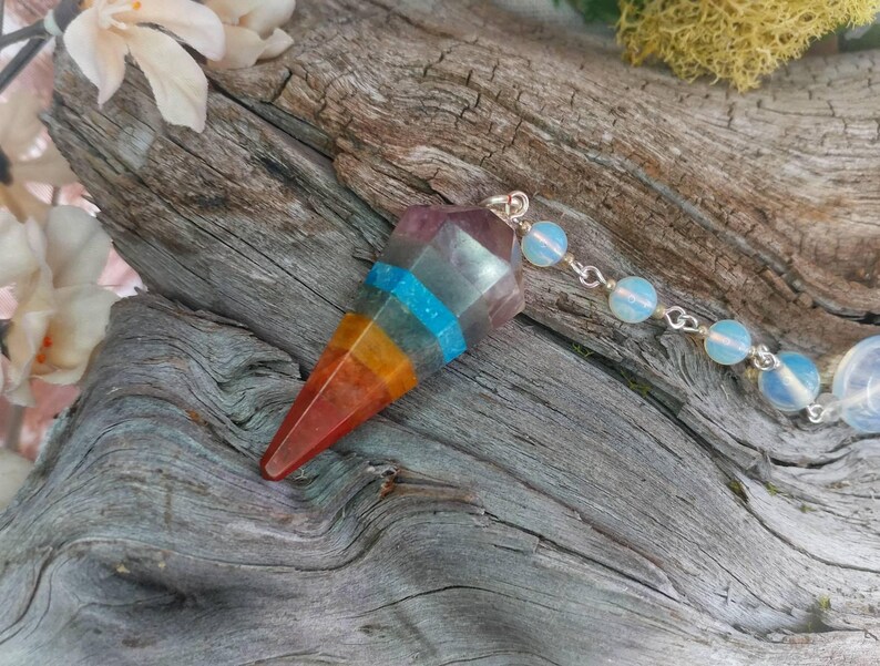 Puede incluir: Un p&eacute;ndulo de cristal arco&iacute;ris con una cadena de plata y cuentas de &oacute;palo blanco. El p&eacute;ndulo es un cristal puntiagudo con capas de rojo, naranja, amarillo, azul y morado.