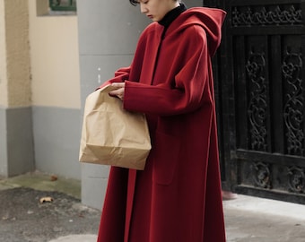 May include: A long, red hooded coat with a button closure and large pockets. The coat is knee-length and has a flowing silhouette. The person is holding a brown paper bag.