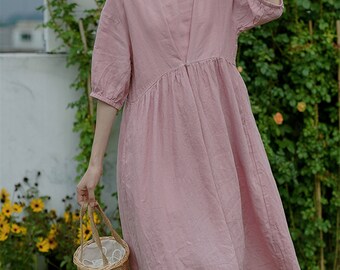 May include: A pink linen dress with a round neckline and 3/4 sleeves. The dress has a gathered skirt and a button closure at the neckline. The model is wearing a white headband and holding a wicker basket.