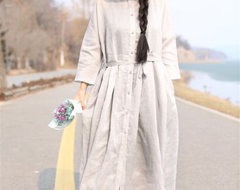 May include: A long, light gray linen dress with a button-up front and a tie belt. The dress has a relaxed fit and a slightly flared skirt. The model is wearing a black hat and white sandals.