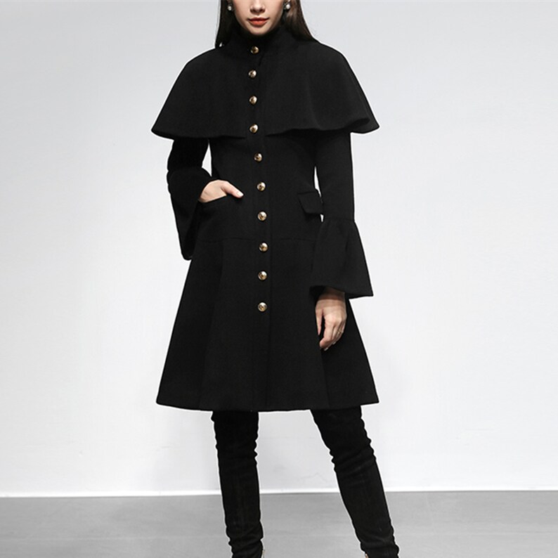 May include: A black coat with a cape-like overlay and flared sleeves. The coat features a high collar and is fastened with gold-colored buttons. The coat has a flared skirt and pockets. The model is wearing black boots.