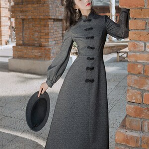 May include: A gray long-sleeved dress with a mandarin collar and a row of black bow knots down the front. The dress has a slightly flared skirt and is made of a textured fabric.