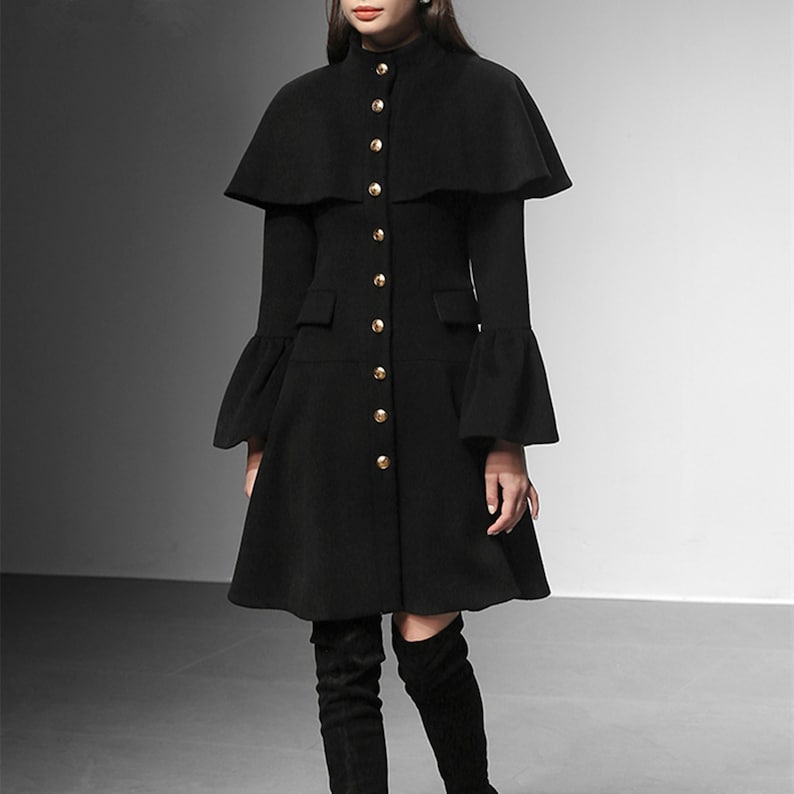 May include: A black coat with a cape-like overlay and gold buttons. The coat features long sleeves with flared cuffs and a flared skirt. The coat has a high collar and is buttoned down the front. The model is wearing black over-the-knee boots.