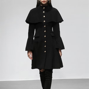 May include: A black coat with a cape-like overlay and flared sleeves. The coat features gold-colored buttons down the front and two pockets. The coat has a high collar and a flared skirt, creating a classic silhouette.