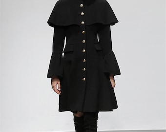 Op de afbeelding: Een zwarte jas met een cape-achtige overlay en uitlopende mouwen. De jas heeft goudkleurige knopen aan de voorkant en twee zakken. De jas heeft een hoge kraag en een uitlopende rok, wat een klassiek silhouet creëert.