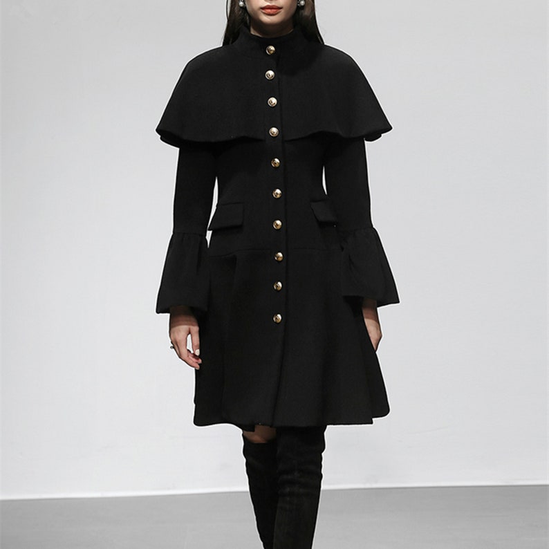 May include: A black coat with a cape-like overlay and flared sleeves. The coat features gold-colored buttons down the front and two pockets. The coat has a high collar and a flared skirt, creating a classic silhouette.