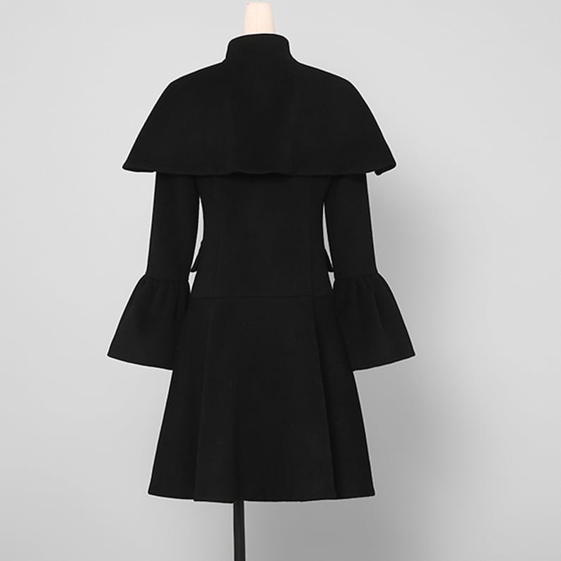 May include: A black coat with a cape-like overlay and flared sleeves. The coat has a high collar and a fitted waist that flares out into a skirt. The coat is displayed on a mannequin against a plain background.