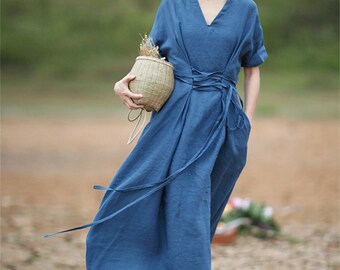 May include: A blue linen dress with a V-neckline and a tie at the waist. The dress has short sleeves and a flowing skirt. The model is wearing a brown hat and holding a woven basket.