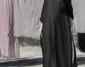 May include: A long, black linen dress with a round neckline and elbow-length sleeves. The dress has side slits and a loose, flowing silhouette. The background features hanging fabric in shades of pink and gray.