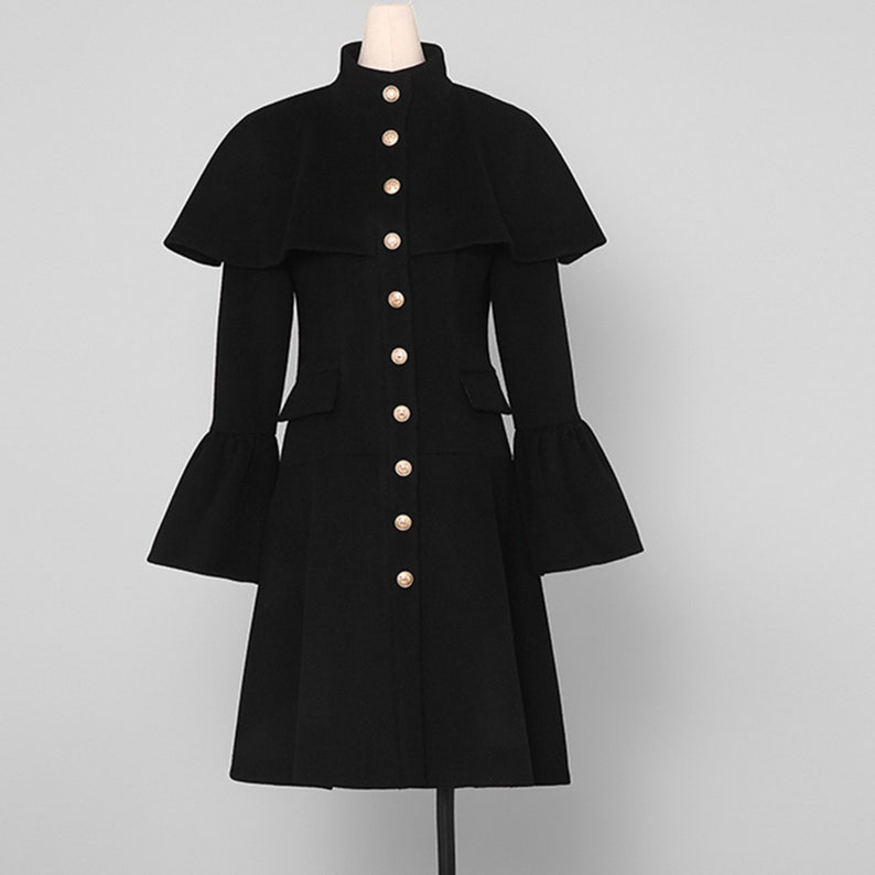 May include: A black coat with a cape-like overlay and flared sleeves. The coat features a high collar and is fastened with gold-colored buttons. The coat has a tailored fit and a knee-length hemline.
