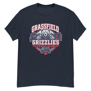 May include: A navy blue short-sleeve t-shirt featuring a graphic of a grizzly bear with headphones and a gaming controller. The text reads GRASSFIELD GRIZZLIES GAMER CLUB in white and red lettering, set against a circular background design.
