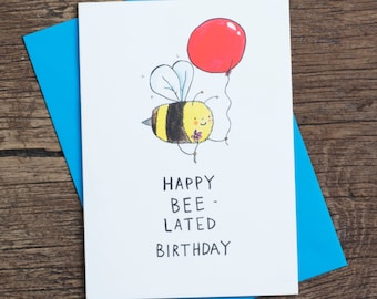 Op de afbeelding: Een witte kaart met een cartoon-bij die een rode ballon vasthoudt. De tekst op de kaart luidt "Happy Bee-Lated Birthday".