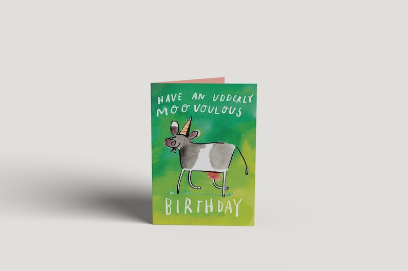 Op de afbeelding: Een groene verjaardagskaart met een cartoonkoe die een feestmuts draagt. De kaart zegt "Have an udderly moo-vellous birthday".