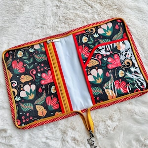 Peut inclure: Une trousse à fermeture éclair à motifs floraux avec un fond noir, une bordure rouge et dorée et une poche transparente. L'étui présente un motif vibrant de fleurs et de feuilles rouges, roses et vertes. Une fermeture éclair jaune avec un charme en forme d'étoile sécurise l'étui.