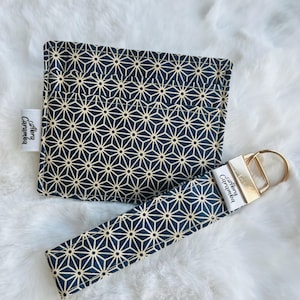 May include: A navy blue and gold geometric pattern features on a small square pouch and a matching wristlet keychain. The keychain has a gold-coloured metal clasp and a white label with the text "Abra Caramba". The items are on a white, fluffy surface.