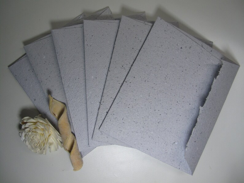 May include: A stack of eight light gray paper envelopes with a speckled texture. The envelopes are arranged in a fan shape.