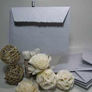 May include: A stack of gray paper envelopes with a textured, speckled surface. The envelopes have a flap on the top and are folded in half. The envelopes are sitting on a table with some decorative items.